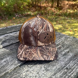 May include: A brown and tan camouflage baseball cap with a brown leather patch featuring a bird and crossed hunting implements. The patch also has the letter "D" in black.