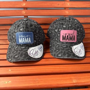 May include: Two black and white leopard print baseball caps with a patch on the front. The left cap has a blue patch with the text "boy mama" and a small heart. The right cap has a pink patch with the text "girl mama" and a small heart.