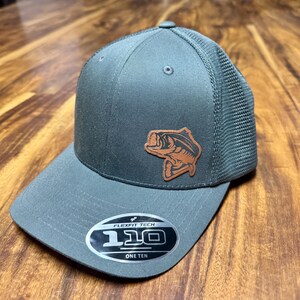 May include: A gray and brown baseball cap with a leather bass fish patch and a Flexfit Tech 110 One Ten label.