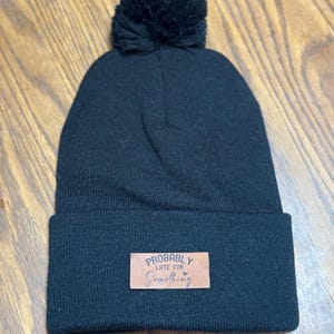 May include: Black knit beanie with a matching pom-pom on top. A rectangular leather patch is sewn on the front, with the words "Probably Late For Something" in white script.
