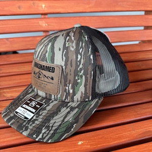 May include: A camouflage trucker hat with a brown leather patch that says "UNASHAMED FOLLOWER OF JESUS CHRIST". The hat is adjustable and has a black mesh back.