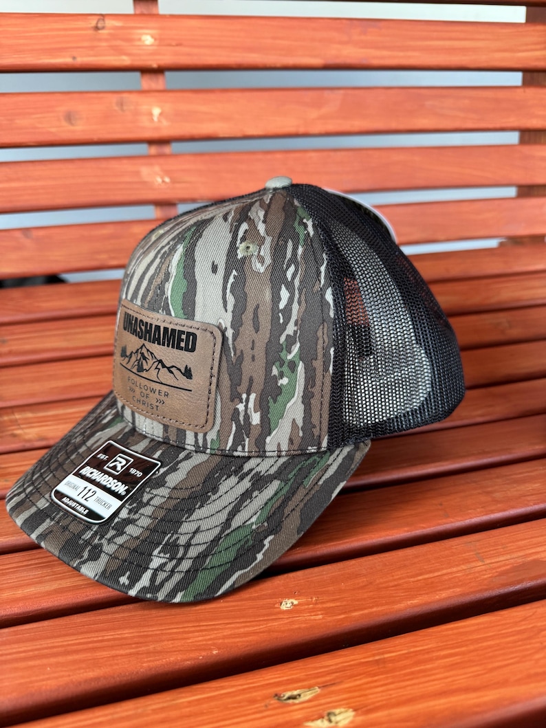 May include: A camouflage trucker hat with a brown leather patch that says "UNASHAMED FOLLOWER OF JESUS CHRIST". The hat is adjustable and has a black mesh back.