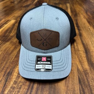 May include: A gray and black trucker hat with a brown leather patch featuring a crossed hunting rifle and a 'G' in the center. The hat has a black mesh back and a 'Richardson' label on the front.