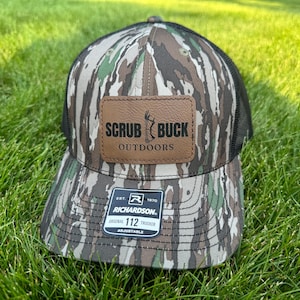 May include: A brown and green camouflage trucker hat with a brown leather patch that says "Scrub Buck Outdoors". The hat is adjustable and has a black mesh back.