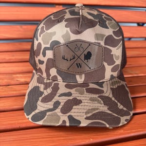 May include: A camouflage trucker hat with a brown leather patch featuring a turkey, antlers, fishing hooks, and the letter "W".