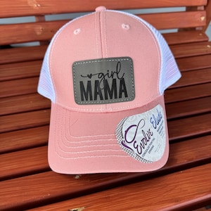 May include: A pink baseball cap with a white mesh back. The cap features a rectangular patch with the words "girl MAMA" in black lettering. A tag on the brim reads "Evolve Elude".