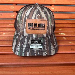 May include: Camouflage trucker hat with a brown leather patch that reads "DAD OF GIRLS #OUTNUMBERED". The hat has a black mesh back and a Richardson 112 Trucker tag.
