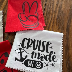May include: Two red microfiber cloths with black designs. One cloth has a Mickey Mouse hand with two fingers up. The other cloth has a white anchor and the text "Cruise Mode On" with a power button symbol.