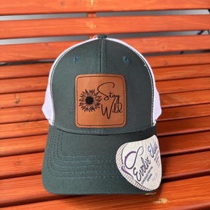 Pu&ograve; includere: Un cappello da camionista verde scuro e bianco con una toppa in pelle marrone che dice "Stay Wild" con una grafica di girasole. Il cappello ha una parte posteriore in rete bianca e un'apertura per la coda di cavallo nascosta.