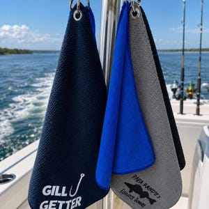 May include: Two textured microfiber towels hanging from metal clips. The left towel is navy blue with white text reading GILL GETTER and a fishhook graphic. The right towel is grey with blue trim and black text reading PAPA RANDY'S Fishing Towel.