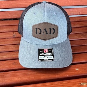 May include: A gray and black trucker hat with a brown leather patch that says "DAD" on it. The hat has a black mesh back and a Richardson 115 label.