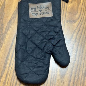 May include: Black quilted oven mitt with a brown patch that reads "my kitchen my rules" in white script. The mitt has a loop for hanging and is designed to protect hands from heat while cooking.