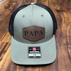 May include: A green and black trucker hat with a brown leather patch that says "PAPA" on it. The hat has a black mesh back and a Richardson 115 Low-Pro Trucker label.