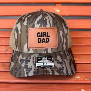 May include: A camouflage trucker hat with a brown leather patch that reads "GIRL DAD" in bold, black letters. The hat has a mesh back and a curved brim. The hat has a Richardson 112 Trucker tag.