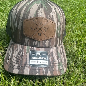 May include: A camouflage trucker hat with a brown leather patch featuring a cross and the letter W. The hat is adjustable and has the text "Richardson" and "112" on the front.