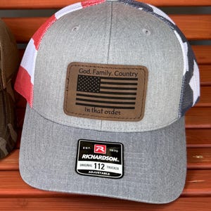 May include: Gray trucker hat with a red, white, and blue mesh back. The hat features a brown leather patch with an American flag and the text "God. Family. Country. In that order."