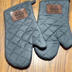 May include: Two gray quilted oven mitts with a diamond pattern. Each mitt has a brown rectangular patch with the words "IN THIS kitchen WE MAKE MESSES AND MEMORIES" in white lettering. The mitts are on a wooden surface.
