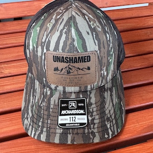 May include: A camouflage trucker hat with a brown leather patch that says "UNASHAMED" and "FOLLOWER OF CHRIST". The hat also has a black mesh back and a black adjustable strap with a brown label that says "EST. 1970 RICHARDSON ORIGINAL 112 TRUCKER ADJUSTABLE".