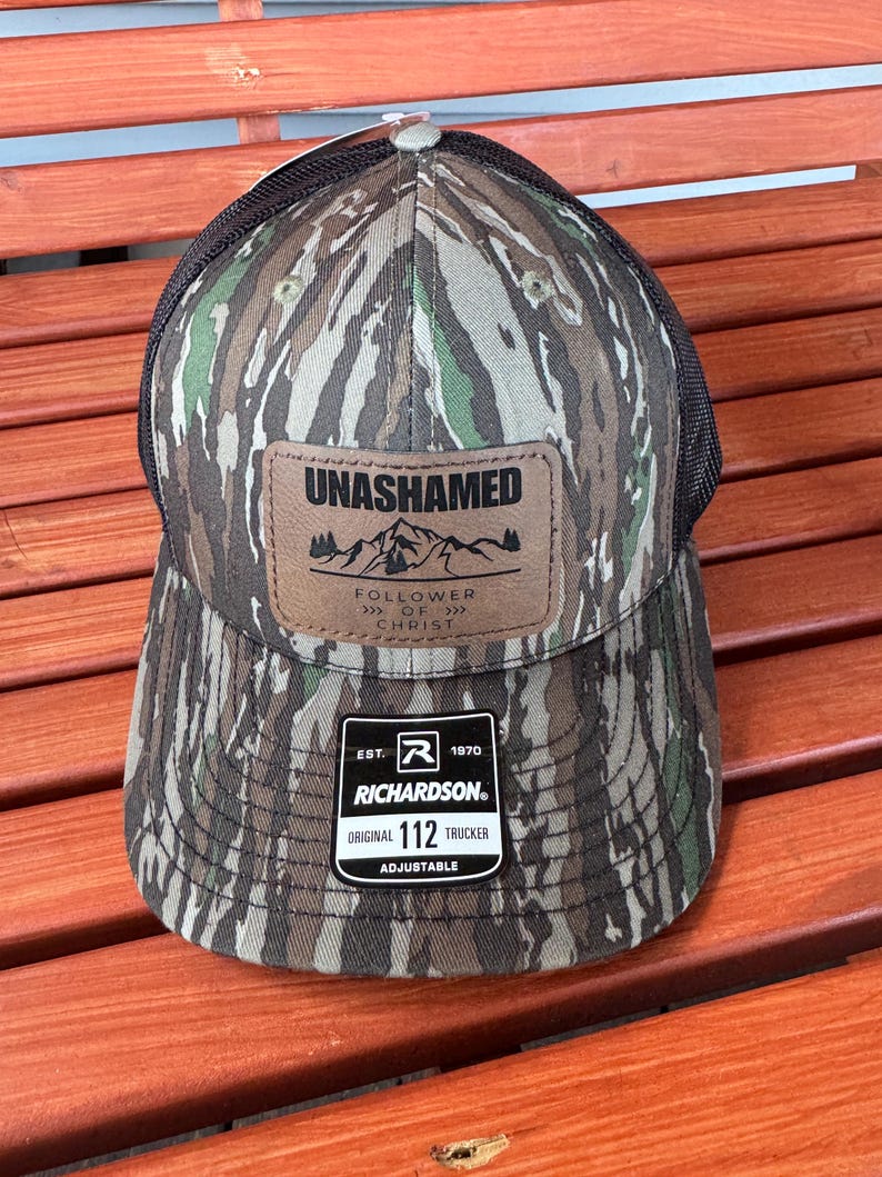May include: A camouflage trucker hat with a brown leather patch that says "UNASHAMED" and "FOLLOWER OF CHRIST". The hat also has a black mesh back and a black adjustable strap with a brown label that says "EST. 1970 RICHARDSON ORIGINAL 112 TRUCKER ADJUSTABLE".