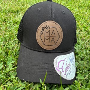 May include: Black trucker hat with a brown leather patch that says "MAMA" in a geometric design. The hat has a mesh back and a curved brim.