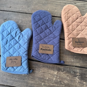 May include: Three quilted oven mitts in blue, navy, and tan. Each mitt has a diamond pattern and a brown leatherette patch with personalized text. The mitts are designed for kitchen use.