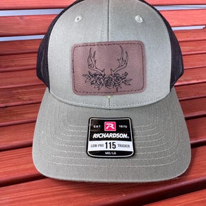 May include: A green and black trucker hat with a brown leather patch featuring a deer antler design with floral accents. The hat has a black mesh back and a Richardson 115 label.