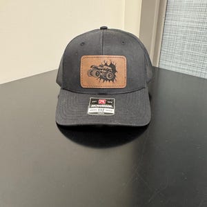 May include: Black trucker hat with a brown leather patch featuring an off-road vehicle design. The hat has a curved brim and a mesh back. The patch has the text "RICHARDSON" and "112 Trucker".