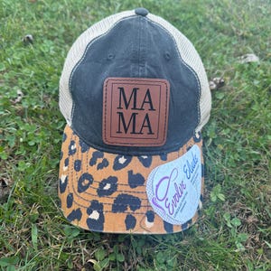 May include: A black and beige trucker hat with a brown leather patch that says "MAMA" in black lettering. The hat has a leopard print brim and a white label that says "Evolve Eluded Concealed Pointers Defining".