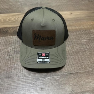 May include: A green and black trucker hat with a brown leather patch that says "Mama" on it. The hat is adjustable and has a black mesh back.