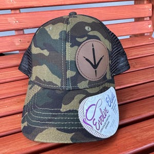 May include: A camouflage trucker hat with a brown leather patch featuring a bird's footprint. The hat has a black mesh back and a label that reads "Evolve Elucidate Concealed  Pontail Opening".