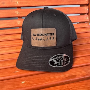 May include: Black Flexfit 110 hat with a mesh back and a curved brim. The hat features a brown leather patch with the text "ALL RACKS MATTER" and deer antler graphics. The hat is sitting on a wooden bench.