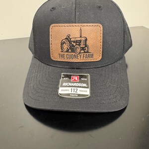 May include: Black trucker hat with a brown leather patch featuring a tractor graphic and the text "THE CUDNEY FARM." The hat has a curved brim and mesh back. A tag reads "RICHARDSON. ORIGINAL 112 TRUCKER YOUTH."