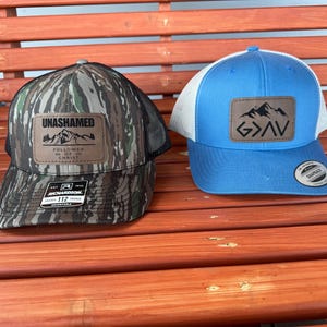 May include: Two trucker hats, one with a camouflage pattern and the words "UNASHAMED" and "FOLLOWER OF MY CHRIST" on a brown patch, and the other with a light blue crown and a brown patch with the words "GOD" and mountains on it.