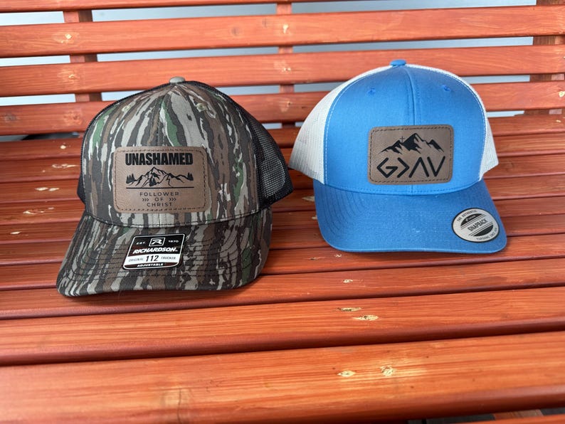May include: Two trucker hats, one with a camouflage pattern and the words "UNASHAMED" and "FOLLOWER OF MY CHRIST" on a brown patch, and the other with a light blue crown and a brown patch with the words "GOD" and mountains on it.