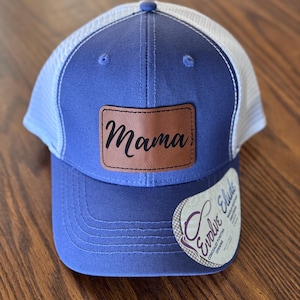 May include: A blue trucker hat with a white mesh back. The hat features a brown leather patch with the word "Mama" in black script. A tag with the words "Evolve Elude" is attached to the side.