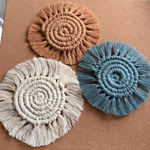 May include: Three macrame coasters in shades of beige, brown, and teal. Each coaster features a spiral center and fringed edges. The coasters are round and appear to be handmade.