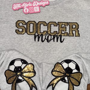 May include: A light gray crewneck sweatshirt featuring the text "SOCCER mom" in gold glitter and black script lettering. Each sleeve cuff displays a black and white soccer ball graphic adorned with a large, gold glitter sequin bow.