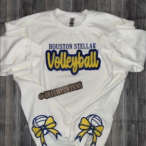 May include: White sweatshirt with the text "HOUSTON STELLAR Volleyball" in blue and yellow lettering. The text "@HILLGIRLSDESIGNS" is below the main text. The sleeves have a yellow bow with a blue volleyball on each cuff.