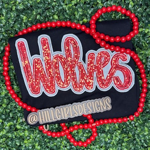 May include: Black t-shirt with the word "Wolves" in red sequined letters with a white outline. A red beaded necklace is draped around the shirt. The text "@HILLGIRLSDESIGNS" is printed on a wooden tag.
