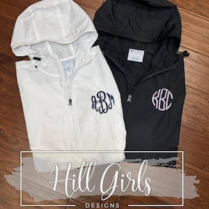 May include: A white and a black Champion brand windbreaker jacket, each featuring a hood with drawstrings, a front zipper, and a personalized monogram embroidered on the left chest. The text Hill Girls DESIGNS is overlaid on the image.