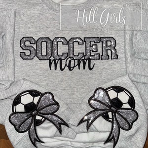 May include: A light gray crewneck sweatshirt featuring the text SOCCER mom in black and silver glitter lettering. Two embroidered patches are positioned at the bottom, each depicting a black and white soccer ball adorned with a silver glitter bow.