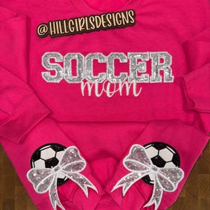 May include: A bright pink long-sleeve sweatshirt featuring the text @HILLGIRLSDESIGNS in black, with the word SOCCER in silver glitter block letters and mom in white script. Each sleeve cuff displays a black and white soccer ball with a silver sequin bow.