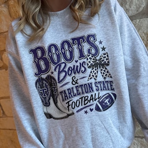 May include: A light gray crewneck sweatshirt featuring a graphic print with the text "BOOTS Bows & TARLETON STATE FOOTBALL". The design includes a pair of white and purple cowboy boots, a leopard print bow, and a purple football with a white T logo.