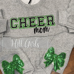 May include: A heather gray crewneck sweatshirt featuring the word CHEER in black and green block letters, with the word mom in black script below. Each sleeve cuff is decorated with a large, bright green sequined bow appliqué.