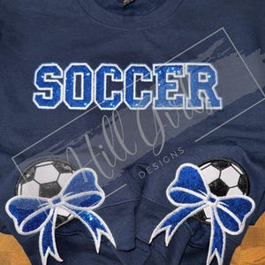 May include: A navy blue crewneck sweatshirt featuring the word SOCCER in blue sequined block letters across the chest. The sleeves display circular patches of black and white soccer balls adorned with large, bright blue sequined bows.