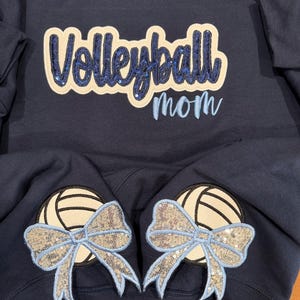 Puede incluir: Una sudadera azul marino con la palabra Volleyball en letras de lentejuelas azul oscuro sobre un parche crema, con la palabra mom bordada en azul claro debajo. Dos parches de voleibol crema con lazos de lentejuelas azul claro en las mangas.