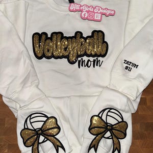 Puede incluir: Una sudadera blanca de manga larga con la palabra Volleyball en purpurina dorada con borde negro, y la palabra mom en letra cursiva negra. Las mangas muestran lazos de purpurina dorada con gr&aacute;ficos de voleibol y el texto TATUM #11.