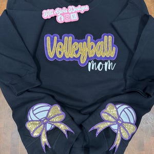 Puede incluir: Una sudadera negra de manga larga con el texto Volleyball mom en purpurina dorada y letra morada. Dos gr&aacute;ficos de voleibol con lazos de purpurina dorada est&aacute;n en las mangas. Un logotipo rosa de Hill Girls Designs se encuentra en la parte superior.