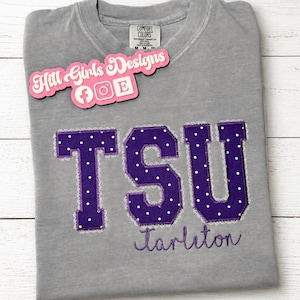 May include: A grey crewneck t-shirt featuring the letters TSU in purple fabric with white polka dots and a frayed edge. Below the letters, the word Tarleton is embroidered in purple script. A pink logo tag reads Hill Girls Designs.