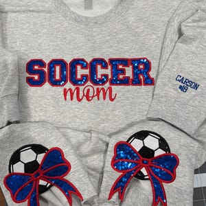 May include: A light gray crewneck sweatshirt featuring the text SOCCER in blue sequin letters with red outlines and mom in red script. The sleeves display soccer ball graphics with blue sequin bows and red outlines. CARSON #18 is embroidered in blue.